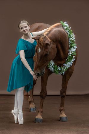 Ballet dancer in turquoise dress poses gracefully with adorned horse.