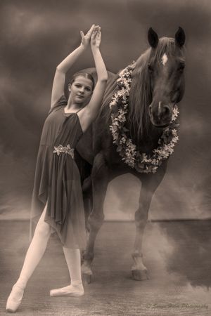 Sepia toned portrait of ballerina in flowing dress performing alongside decorated horse against moody sky.