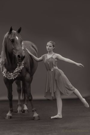 Monochrome image of dancer in arabesque position next to horse wearing floral necklace.