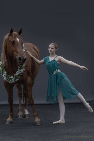 Ballet dancer in teal dress strikes elegant pose beside brown horse adorned with floral garland.