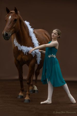 Dancer in teal dress poses with horse wearing white garland against dark studio background.