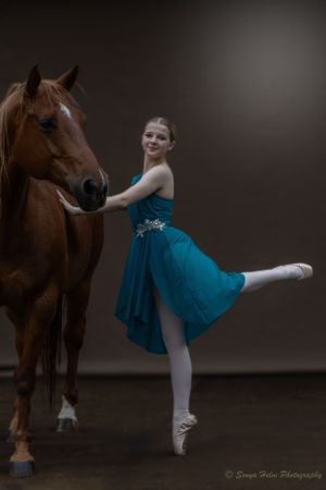 Graceful ballerina in turquoise dress performs arabesque while holding onto horse in studio.