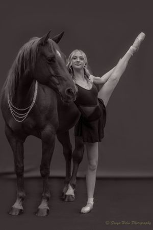 Black and white portrait of ballet dancer performing développé while touching horse's neck.