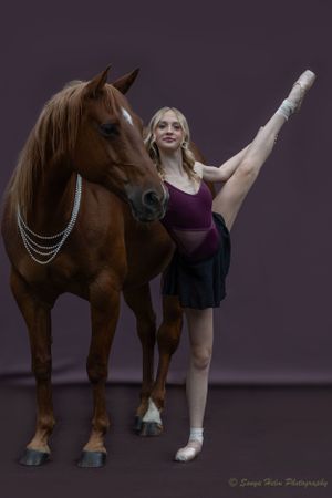 Dancer in purple leotard executes high leg extension while posing with brown horse wearing chains.