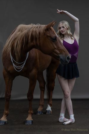 A dancer in purple dress strikes a graceful pose beside a chestnut horse wearing a chain necklace.