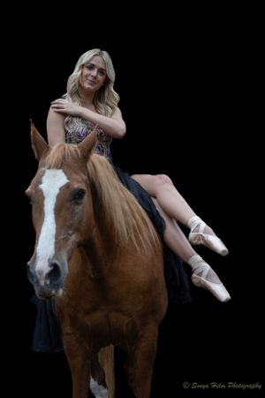 A ballet dancer in a black dress sits gracefully atop a chestnut horse with white markings.