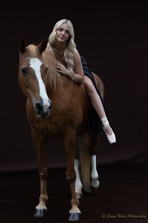 A dancer in ballet attire poses on a chestnut horse against a dark background.