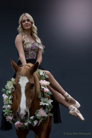 A dancer sits sideways on a horse wearing a floral garland in an artistic portrait.
