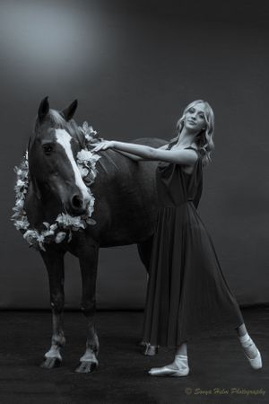 A black and white photo of a dancer in pointe shoes posing with a flower-adorned horse.