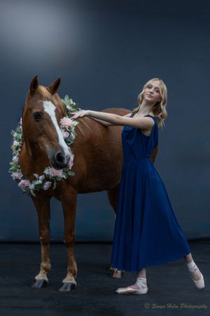 A ballerina in a flowing blue dress poses gracefully next to a brown horse wearing a floral garland.