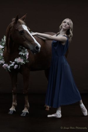 A dancer in blue dress and pointe shoes stands beside a brown horse adorned with flowers.