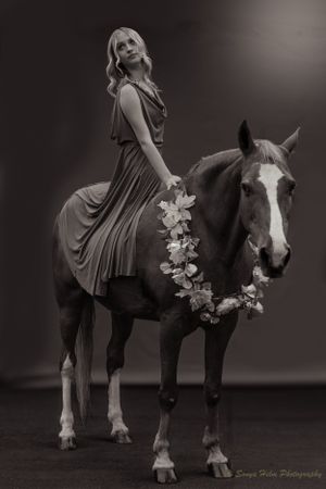 A monochrome portrait of a rider in a dress mounted on a horse wearing a floral wreath.