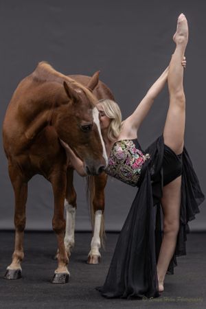 A dancer in a flowing black dress demonstrates extreme flexibility while interacting with a chestnut horse.