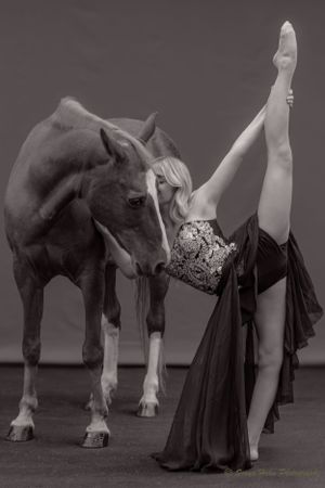 Dramatic black and white photo of a dancer's vertical split pose next to a horse in moody lighting.