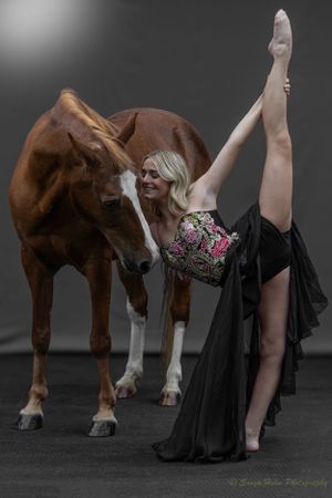 A dancer in elegant costume performs a dramatic leg extension while sharing a moment with a horse.