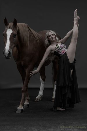 Artistic portrait of a dancer's vertical split pose alongside a bay horse in moody lighting.