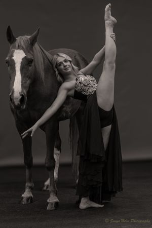 Monochrome image of a dancer performing an arabesque pose next to a horse with white facial markings.