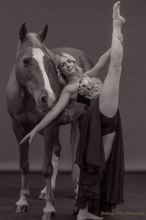 Black and white portrait of dancer executing dramatic leg extension pose beside majestic horse.