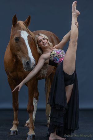 Athletic dancer performs high leg extension pose alongside brown horse with white blaze in dramatic studio setting.