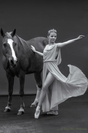 A dancer in a flowing white dress and tiara strikes an elegant pose beside a dark horse in black and white.