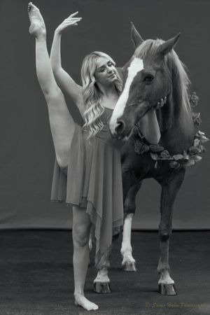 Graceful dancer in flowing dress performs elegant leg extension pose beside chestnut horse in dramatic black and white photo.