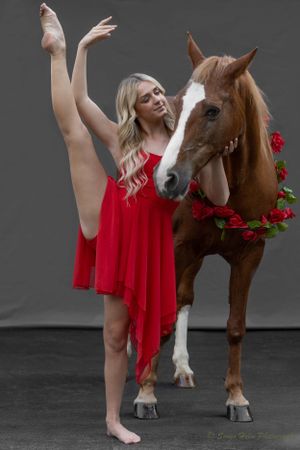 A dancer in red performs a high leg extension pose beside a horse adorned with roses.