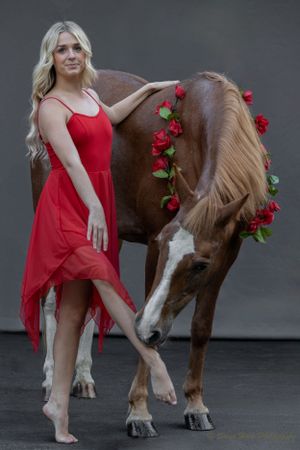 Red-dressed dancer poses beside brown horse adorned with roses in artistic portrait.