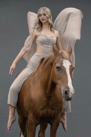 Dancer in beige outfit with flowing fabric poses gracefully on brown and white horse.