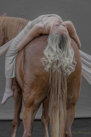 Artistic portrait of dancer in pale dress reclining on brown horse's back.