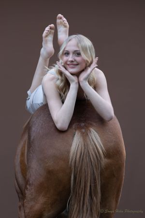 Playful pose of dancer lying on brown horse's back in studio setting.