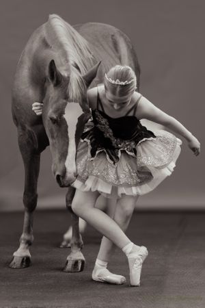 Young dancer in black tutu performs ballet moves beside brown horse in monochrome photo.