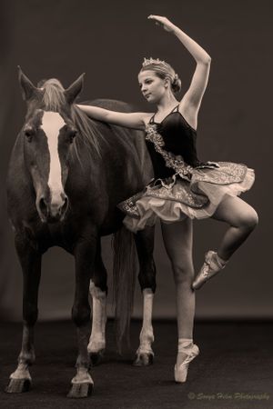 An artistic sepia photo of a ballet dancer posing in a tutu next to a majestic horse.