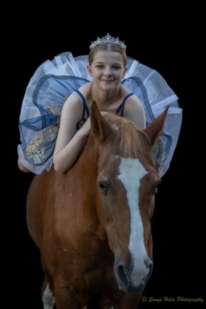 A ballet dancer in a blue tulle tutu and tiara poses with a chestnut horse against black background.