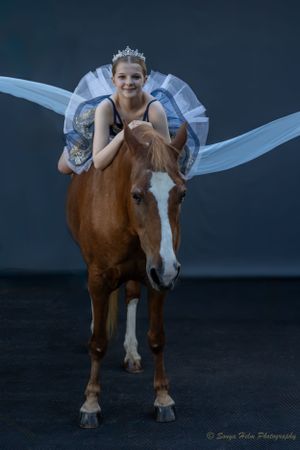 Dancer wearing wings poses with chestnut horse against dark backdrop.
