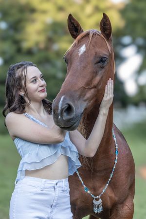 A chestnut colored horse with a white blaze wearing a beaded necklace stands in a grassy outdoor setting.