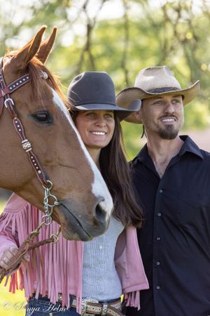 Couple in western attire posing with chestnut horse wearing decorative bridle against blurred natural background.