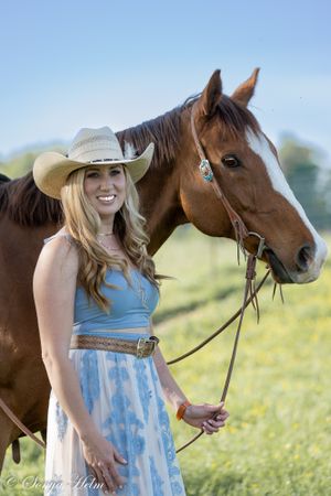 A brown horse and person in a blue dress and cowboy hat pose together in a sunny field.