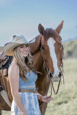 A brown and white horse with a saddle stands next to a person in western attire in a field.