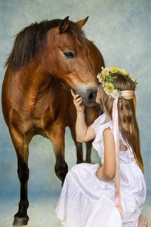 A brown horse nuzzles close to a young figure in a white dress wearing a floral crown against a muted background.