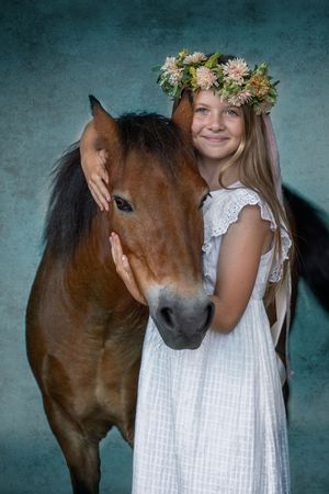 A brown horse nuzzles against a person wearing a white dress and floral crown in an artistic portrait series.