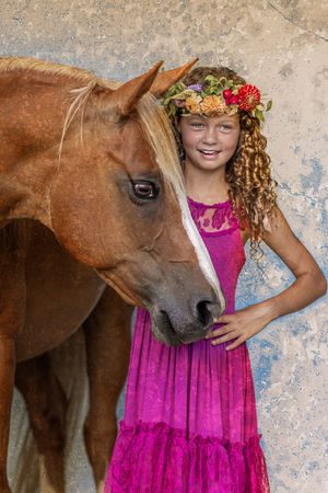 A beautiful brown horse stands beside someone wearing a pink dress and floral crown against a textured wall.