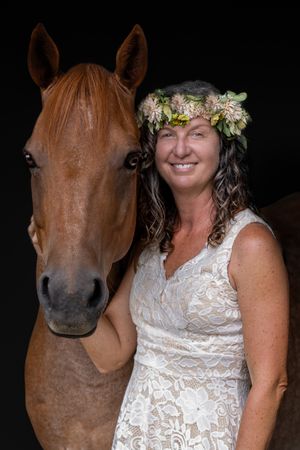 A brown horse stands beside someone wearing a white lace dress and floral crown against a dark background.