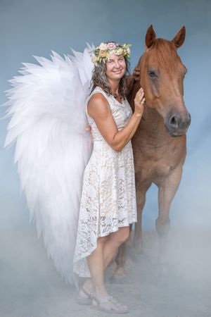 Ethereal photoshoot featuring a white lace dress and brown horse with angel wings in a dreamy blue backdrop.