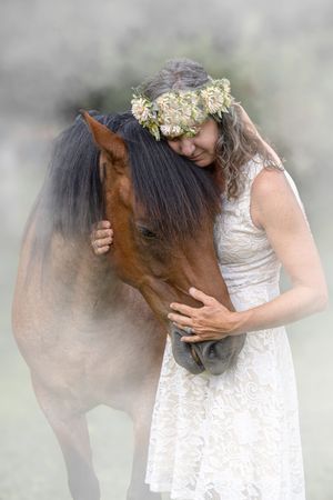An artistic portrait featuring a brown horse and a white lace dress against a soft, misty backdrop.