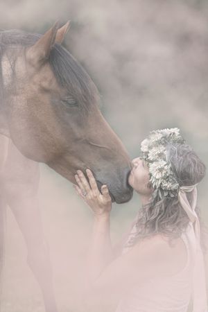 A person and brown horse share a tender interaction with white flowers against a soft, ethereal backdrop.