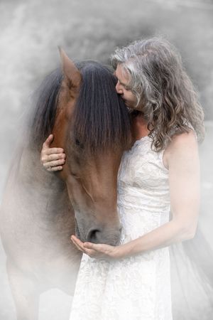 A person in a white dress shares an intimate moment with a dark brown horse against a misty background.