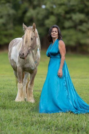 A white horse stands beside someone wearing a flowing turquoise dress in a grassy field during a photo session.
