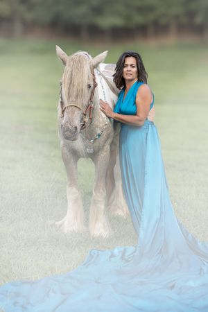 A person in a flowing light blue dress poses with a majestic draft horse in a serene outdoor setting.