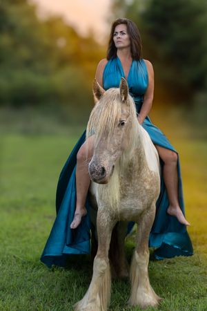 A person in a flowing teal dress sits atop a cream-colored draft horse in a grassy field at sunset.