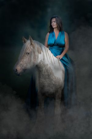 A white horse emerges from misty fog while someone in a teal dress stands beside it in a moody photoshoot.